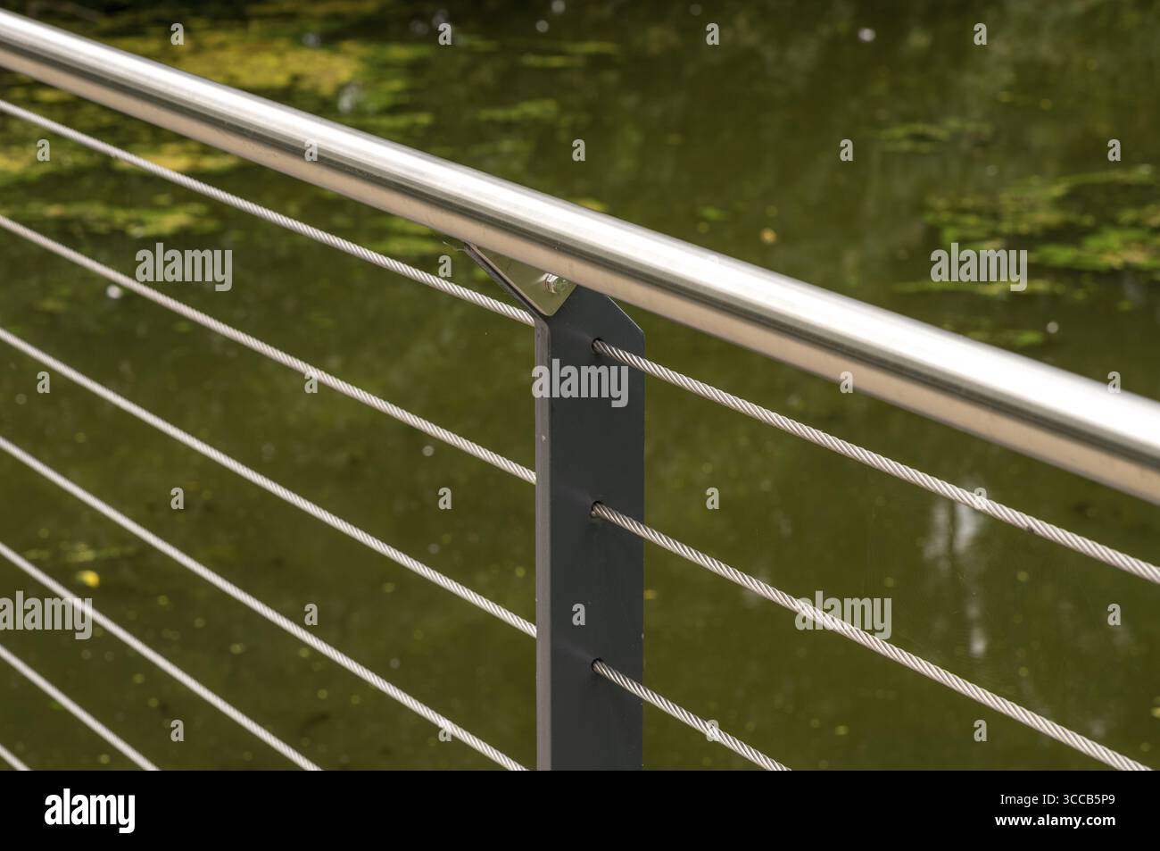 Primo piano di una ringhiera in acciaio con un corpo d'acqua verde sfocato sullo sfondo, creando un senso di tranquillità e un design moderno in una città Foto Stock