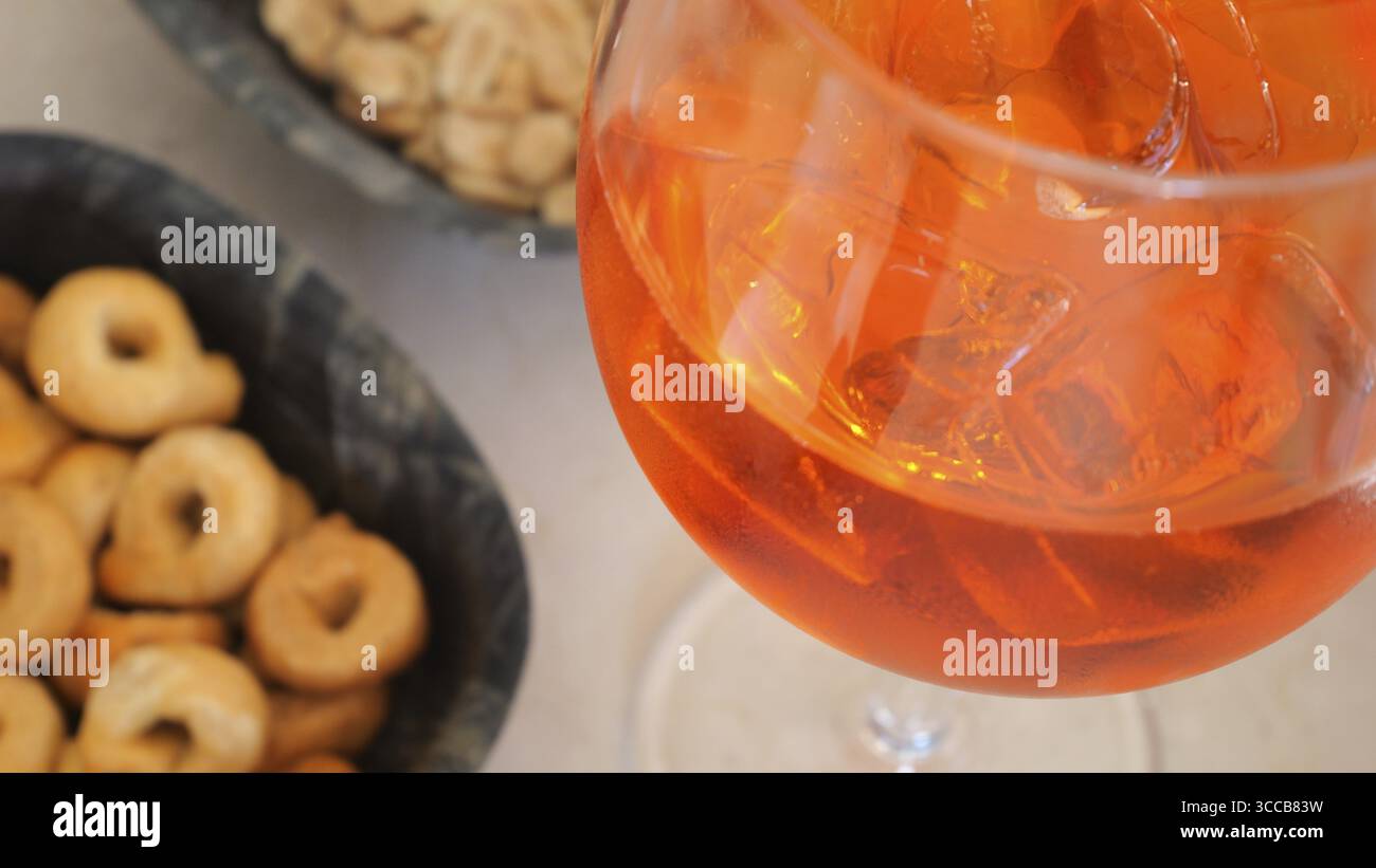Bicchiere da cocktail Spritz riempito di cubetti di ghiaccio, circondato da ciotole di taralli e arachidi, per un delizioso aperitivo italiano perfetto Foto Stock