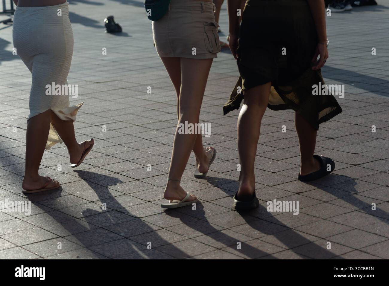 Vista della parte inferiore del corpo delle persone che camminano su una strada asfaltata. Ombre del tardo pomeriggio gettate a terra. Persone che viaggiano. Foto Stock
