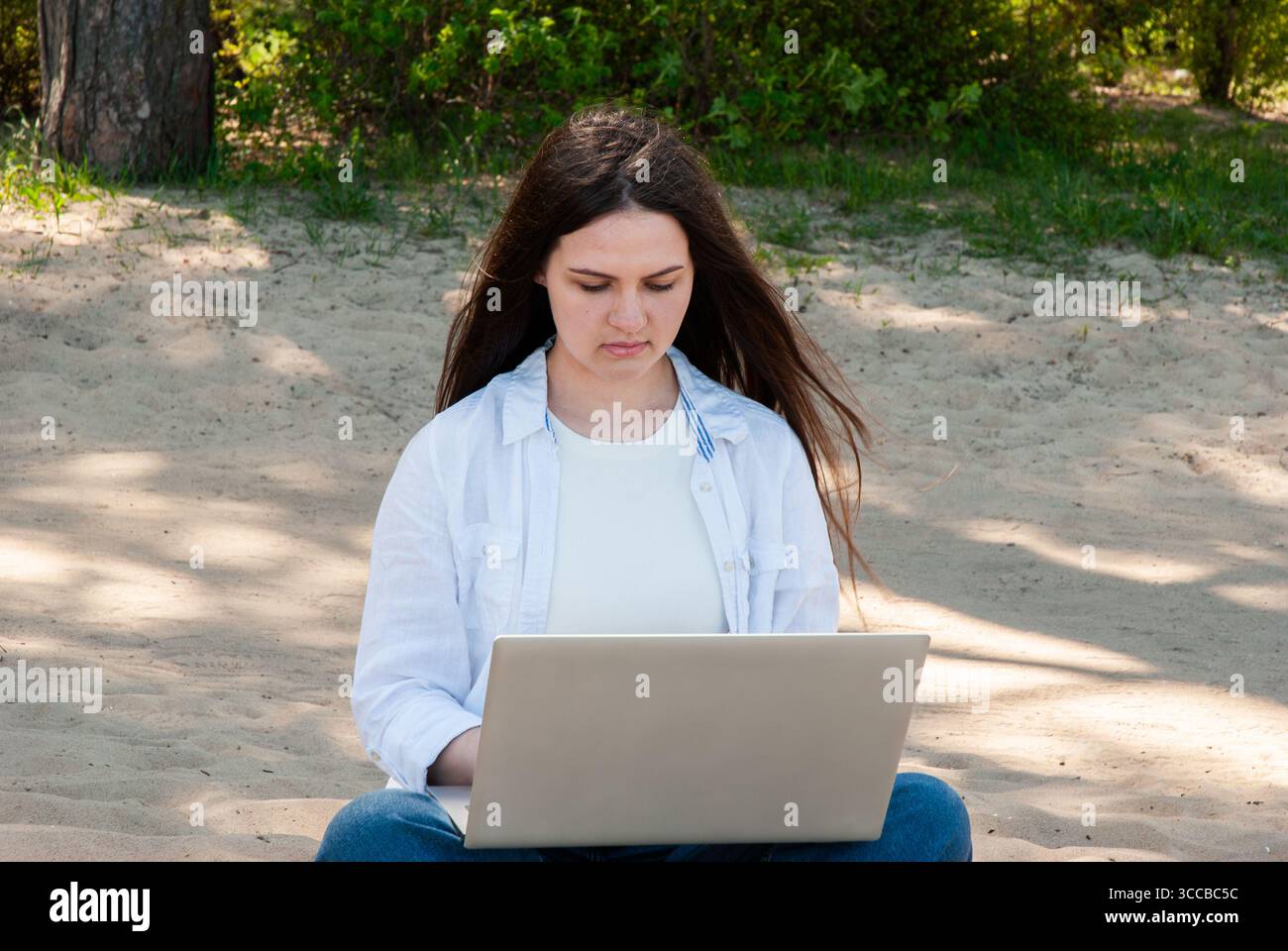 Giovane donna con lunghi capelli castani seduta su un terreno sabbioso in un parco soleggiato, che lavora su un notebook, simboleggia il lavoro a distanza, il freelance e il nomade digitale Foto Stock