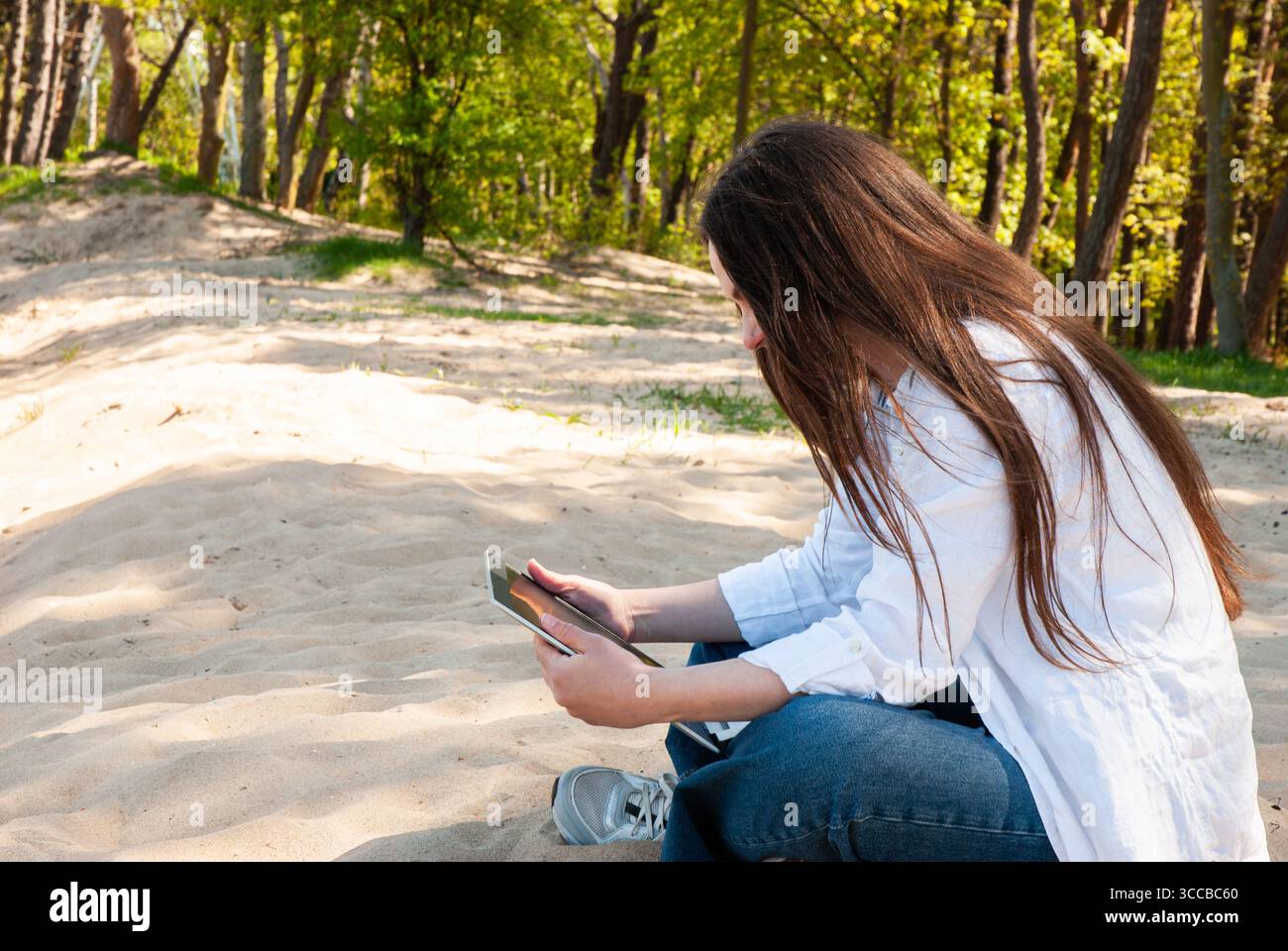 Giovane donna con lunghi capelli castani seduta su un terreno sabbioso in un parco soleggiato, che utilizza un tablet, simboleggia il lavoro a distanza, l'e-learning e il noma digitale Foto Stock