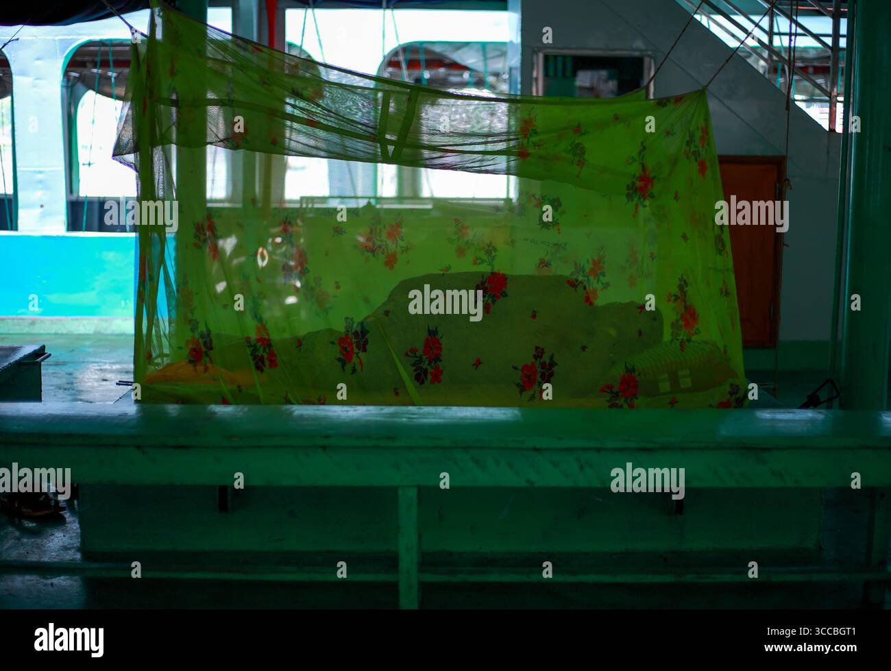 Dhaka, Bangladesh - 17 maggio 2019: Vista di un letto drappeggiato da una zanzariera verde floreale si siede su una piattaforma, gettando ombre nella luce soffusa. Foto Stock