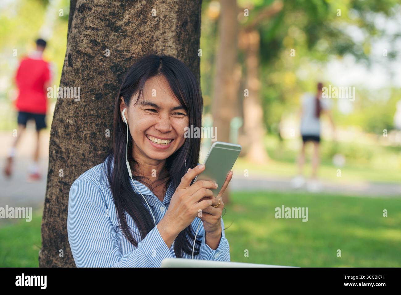 Donna asiatica seduta verde parco giardino utilizzando il telefono ascoltare la musica da smartphone con computer portatile. Felicità donna sorridente, ridendo felice toothy s Foto Stock