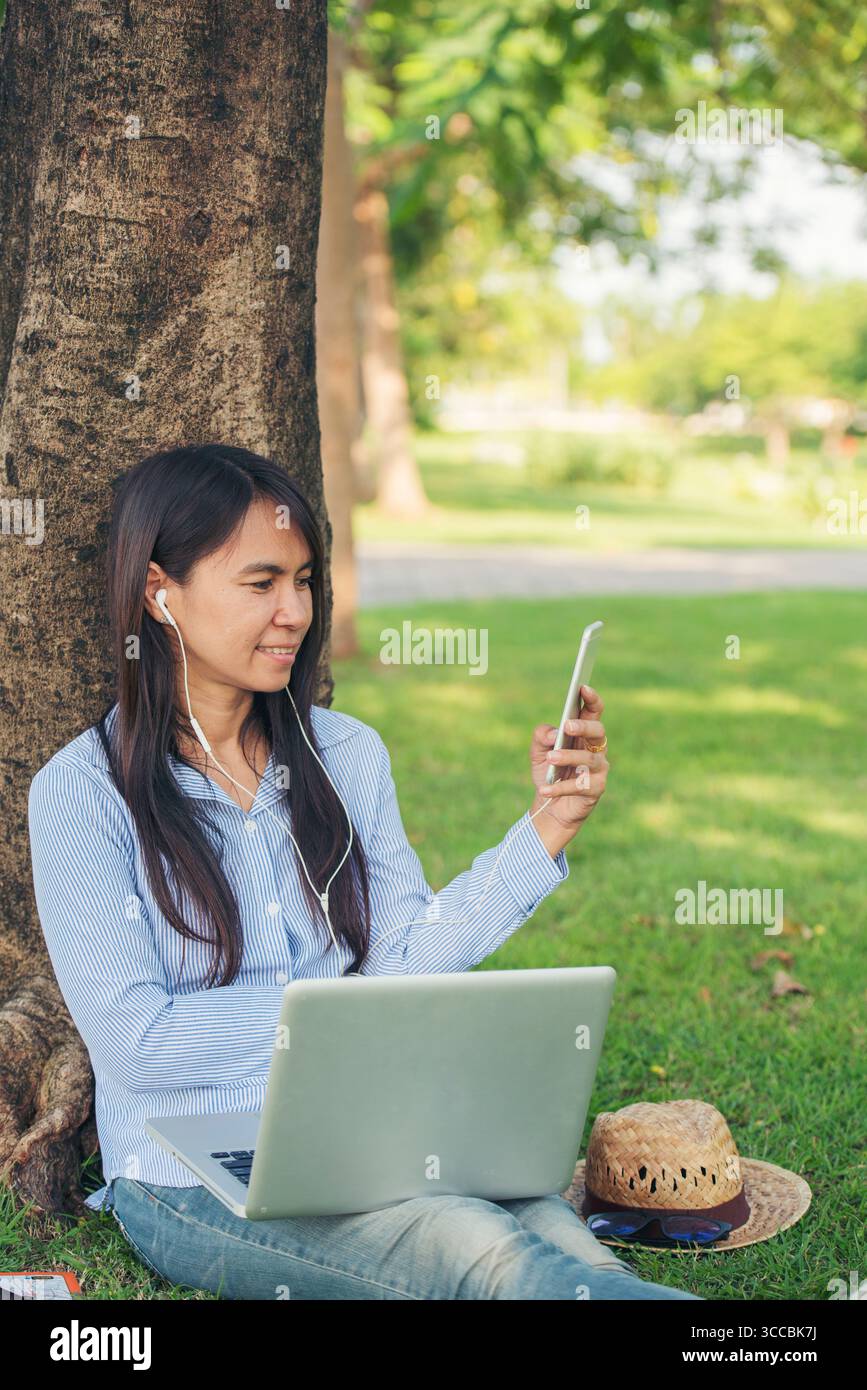 Donna asiatica seduta verde parco giardino utilizzando il telefono ascoltare la musica da smartphone con computer portatile. Felicità donna sorridente, ridendo felice toothy s Foto Stock