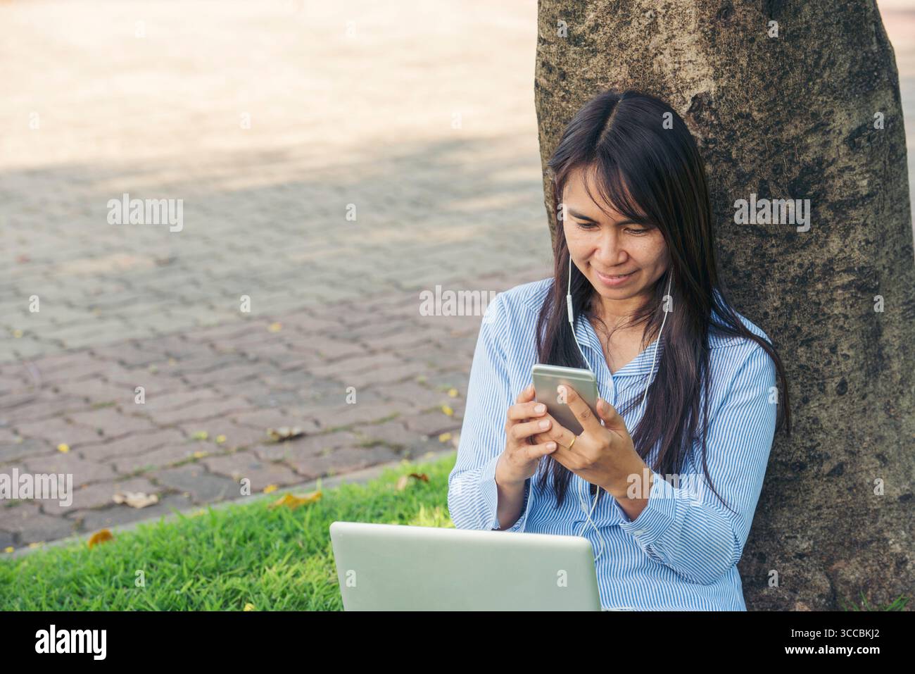 Donna asiatica seduta verde parco giardino utilizzando il telefono ascoltare la musica da smartphone con computer portatile. Felicità donna sorridente, ridendo felice toothy s Foto Stock