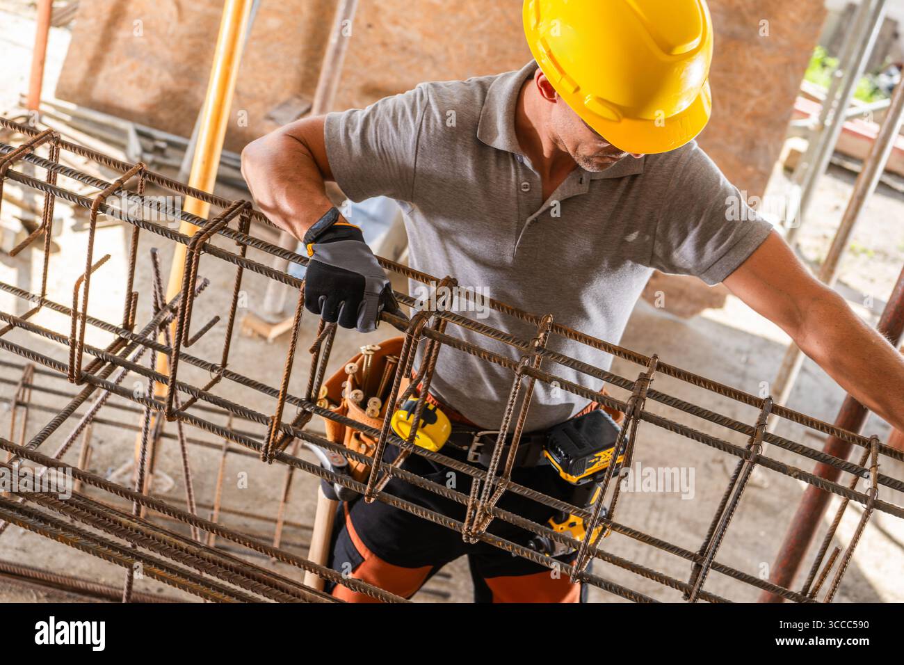 Un operaio edile che indossa un elmetto maneggia con cura i tondini metallici in un cantiere durante il giorno. Gli attrezzi sono visibili sulla sua cintura. Foto Stock