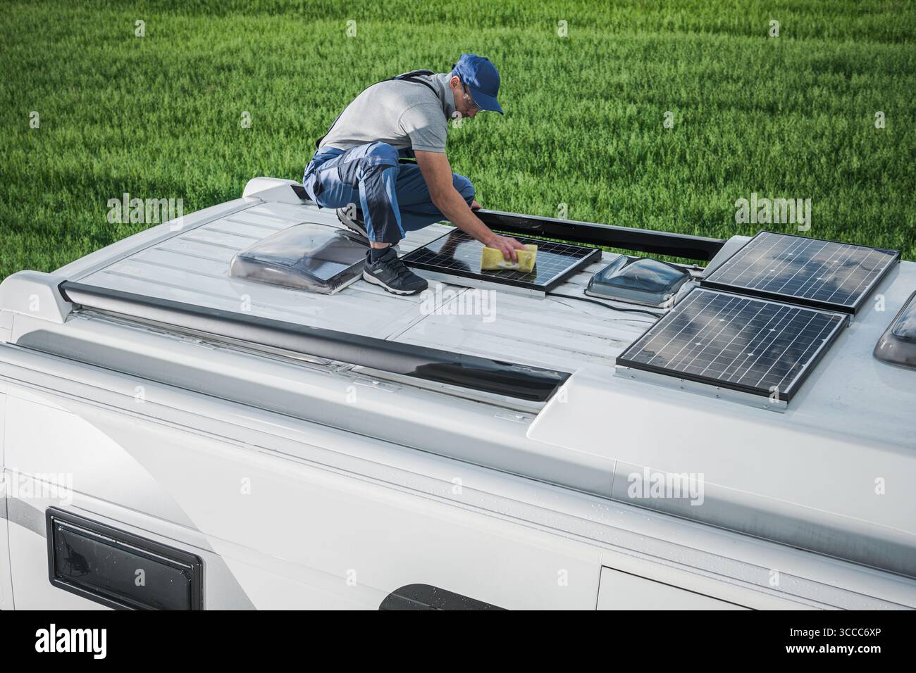 Un uomo ispeziona e pulisce i pannelli solari sul tetto di un veicolo ricreativo in un campo aperto sotto cieli limpidi, garantendo un'efficiente acquisizione di energia. Foto Stock