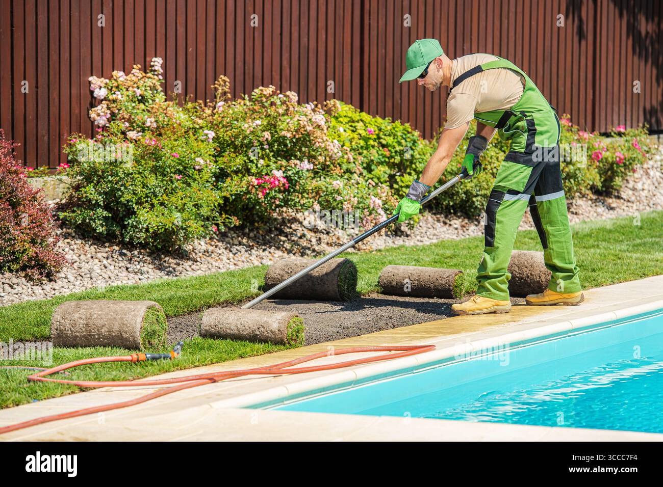 Un giardiniere stende rotoli di sod presso l'area della piscina, concentrandosi sulla creazione di un lussureggiante paesaggio verde con fiori colorati in un ambiente soleggiato all'aperto. Foto Stock