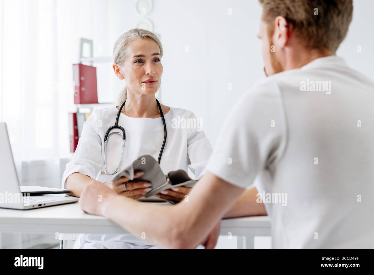 Donna medico in cappotto bianco è consulenza con paziente di sesso maschile in clinica, discutere problemi di salute, con laptop e documenti medici sul tavolo, fosteri Foto Stock