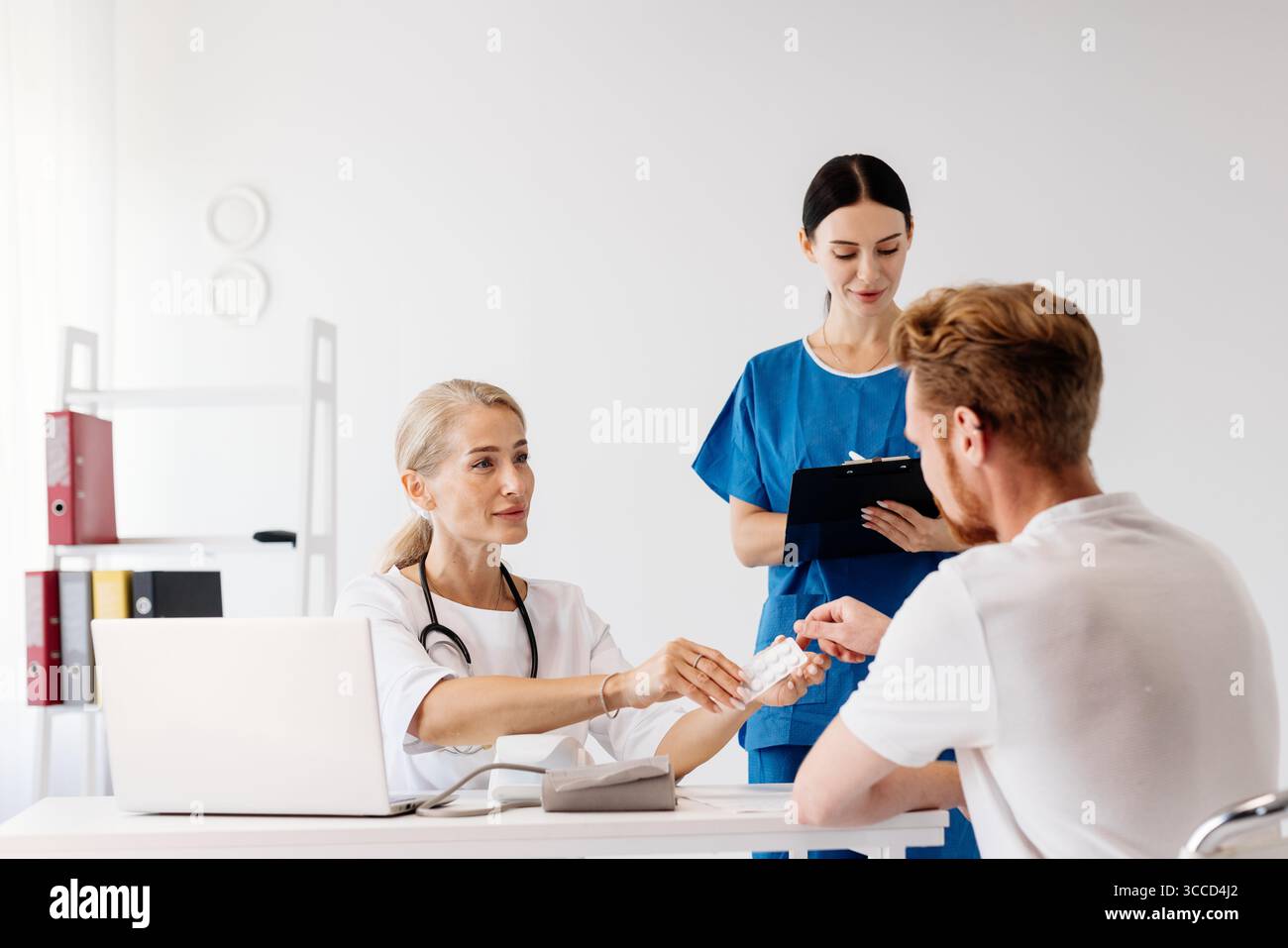 Medico in cappotto bianco è consulenza con paziente di sesso maschile, mentre infermiera assiste con forniture mediche in una clinica moderna, evidenziando la salute professionale Foto Stock
