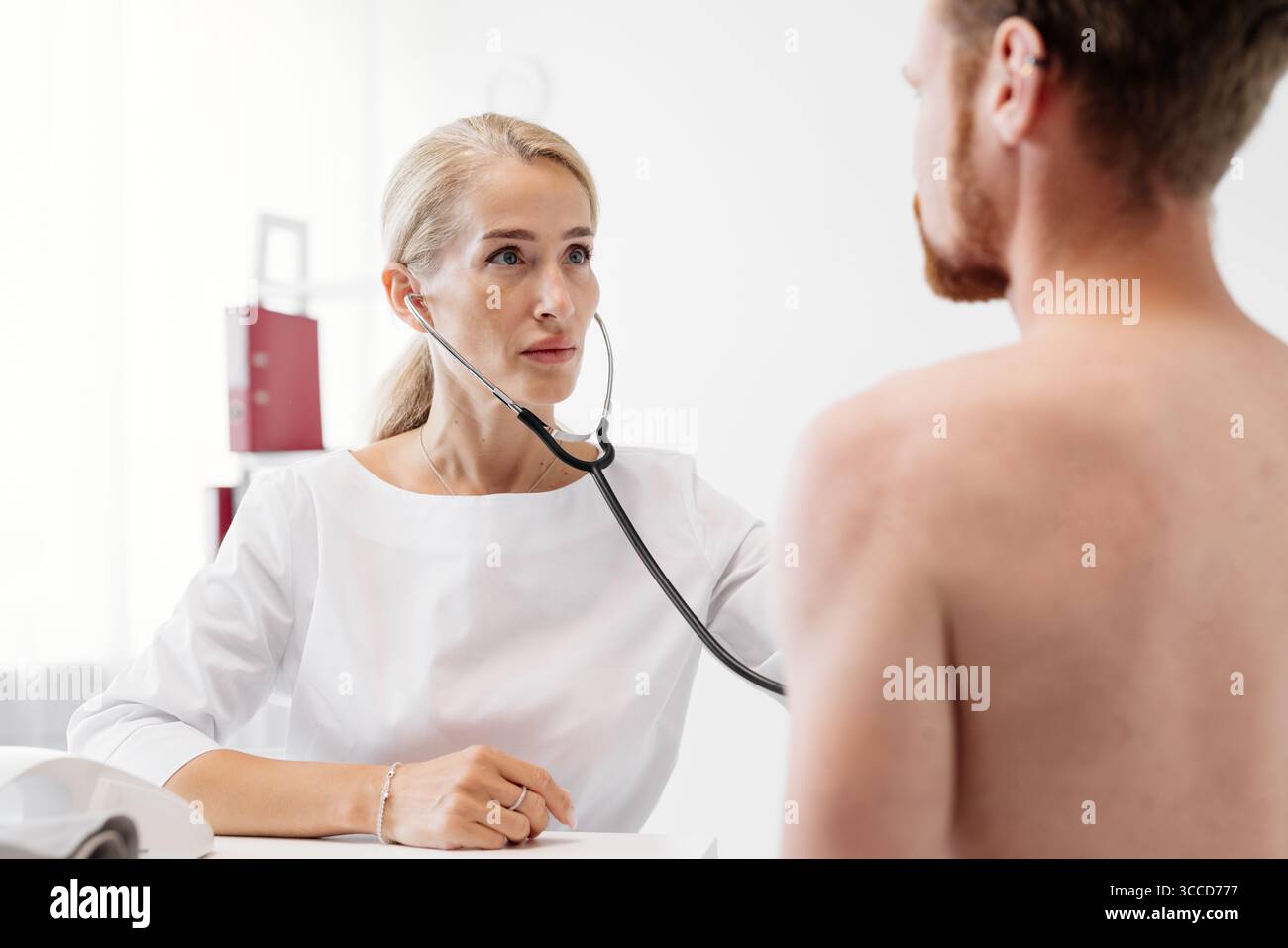 Il medico femminile in camice bianco sta esaminando un paziente di sesso maschile con stetoscopio in clinica luminosa, evidenziando l'importanza della sanità e della cura del paziente Foto Stock