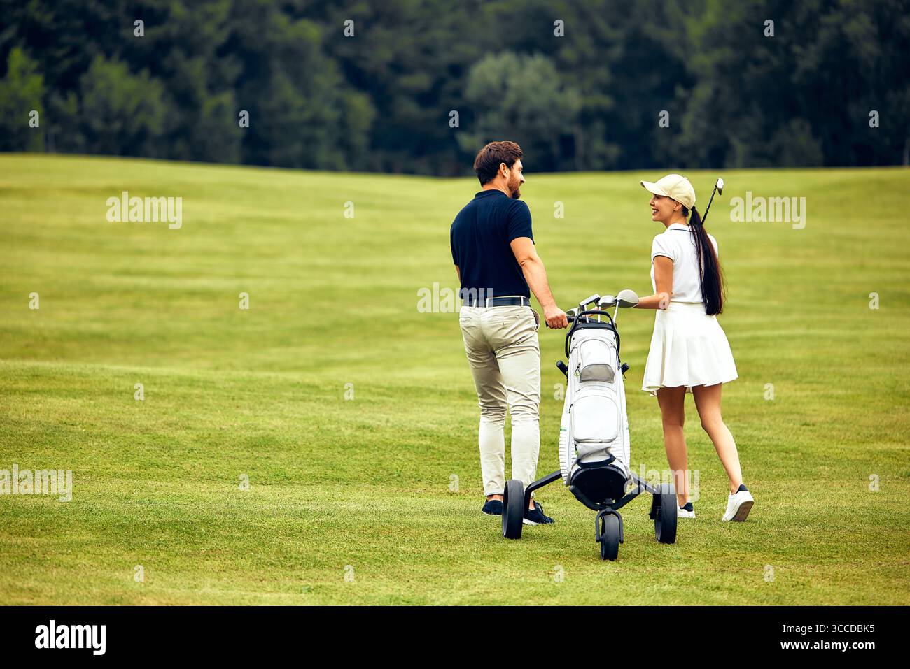 Una coppia elegante cammina piacevolmente mano nella mano su un campo da golf splendidamente curato, trasportando felicemente le loro mazze e godendosi il loro tempo di qualità Foto Stock