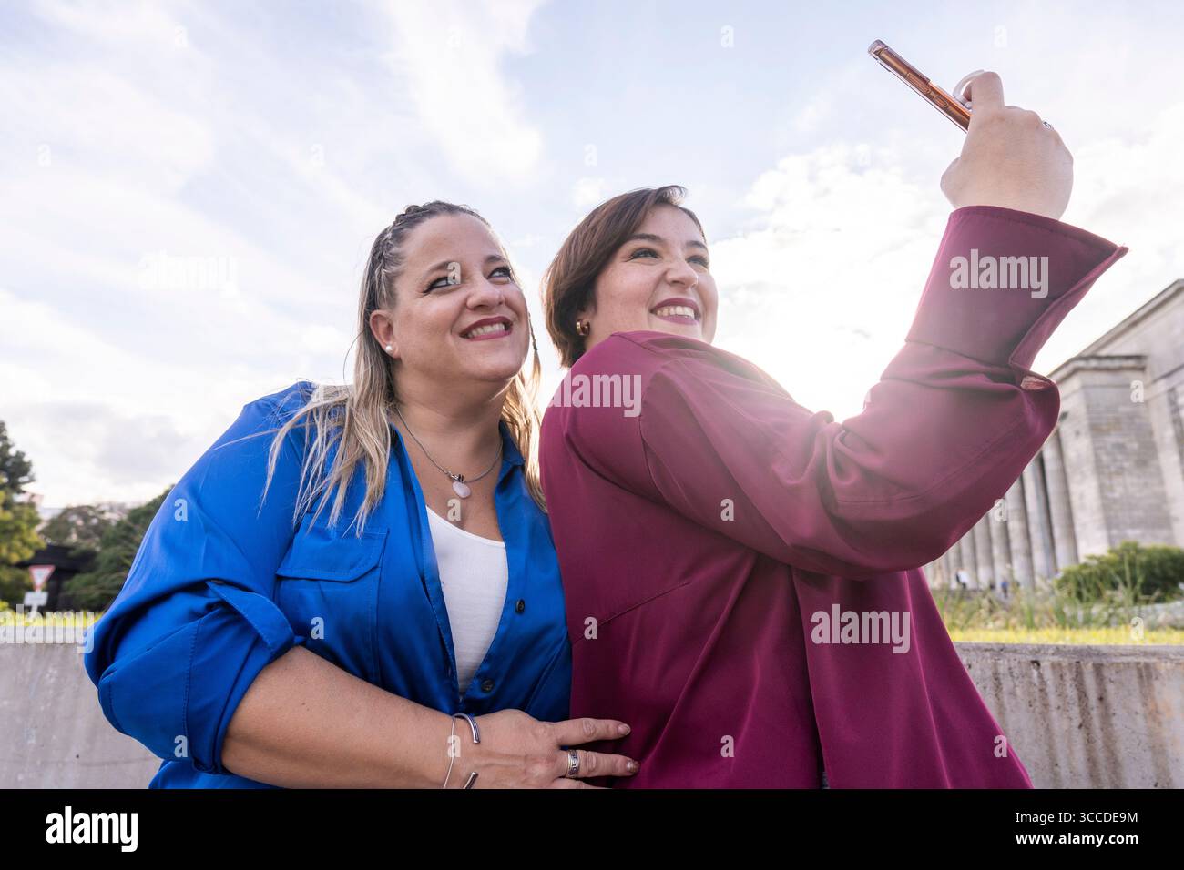 Due amici sorridono e si mettono in posa per un selfie in un parco cittadino, circondato dal verde e dall'architettura storica. Foto Stock
