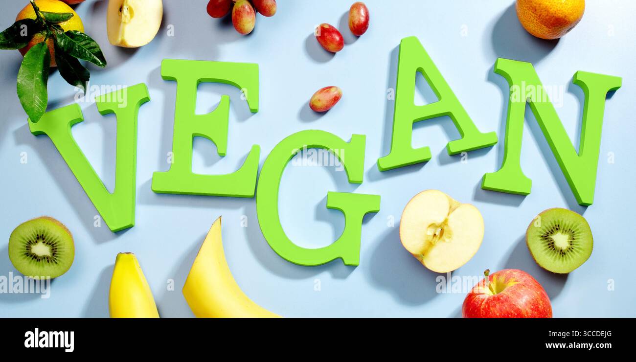 La parola VEGAN in grandi lettere verdi su una superficie blu, circondata da frutta fresca, verdure e una ciotola di lenticchie, dieta vegana piatta Foto Stock