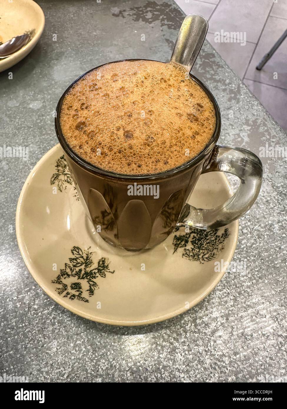 Un caldo e frizzante caffè hailam tradizionale in una tazza di vetro trasparente con cucchiaio, su un piattino decorativo in un rustico caffè kopitiam di kuala lumpur malesia Foto Stock