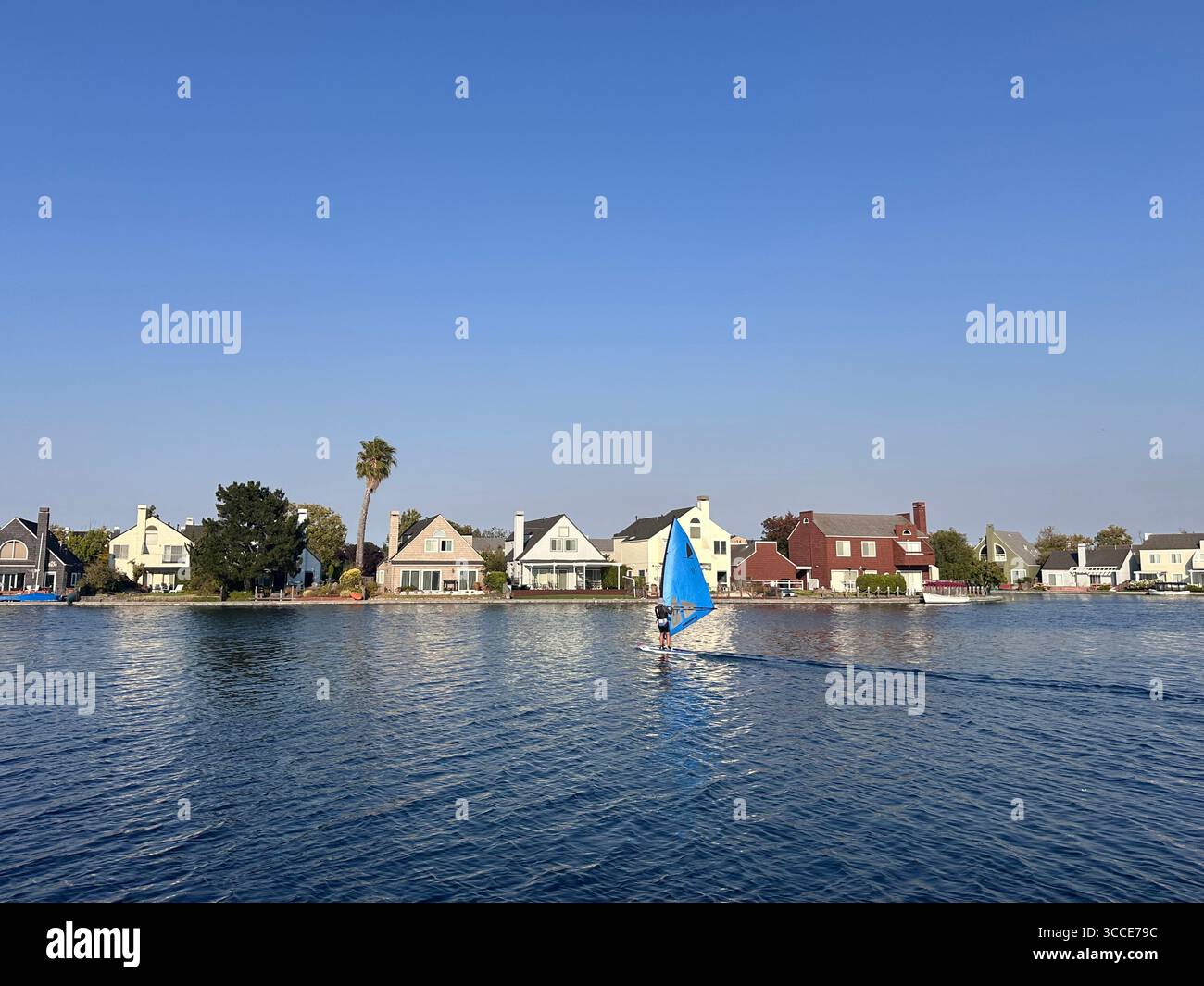 Un windsurfista scivola attraverso un tranquillo lago suburbano di fronte a una fila di case colorate sul lungomare sotto un cielo azzurro - Immagine stock catturata con smartphone