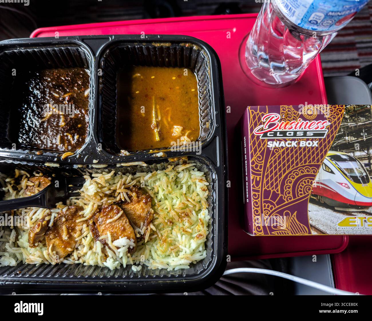 Pasto in treno KTM ETS Business Class con riso, curry e una scatola di spuntini servita su un vassoio rosso con una bottiglia d'acqua di plastica. - Immagine stock catturata con smartphone