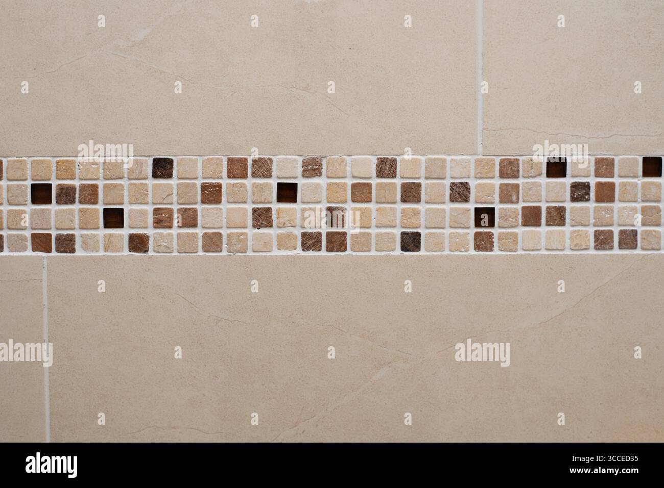 Le piastrelle a mosaico in marrone e creme terrosi creano un bordo decorativo su una parete piastrellata beige, aggiungendo un tocco di stile ai disegni delle pareti interne dei bagni Foto Stock