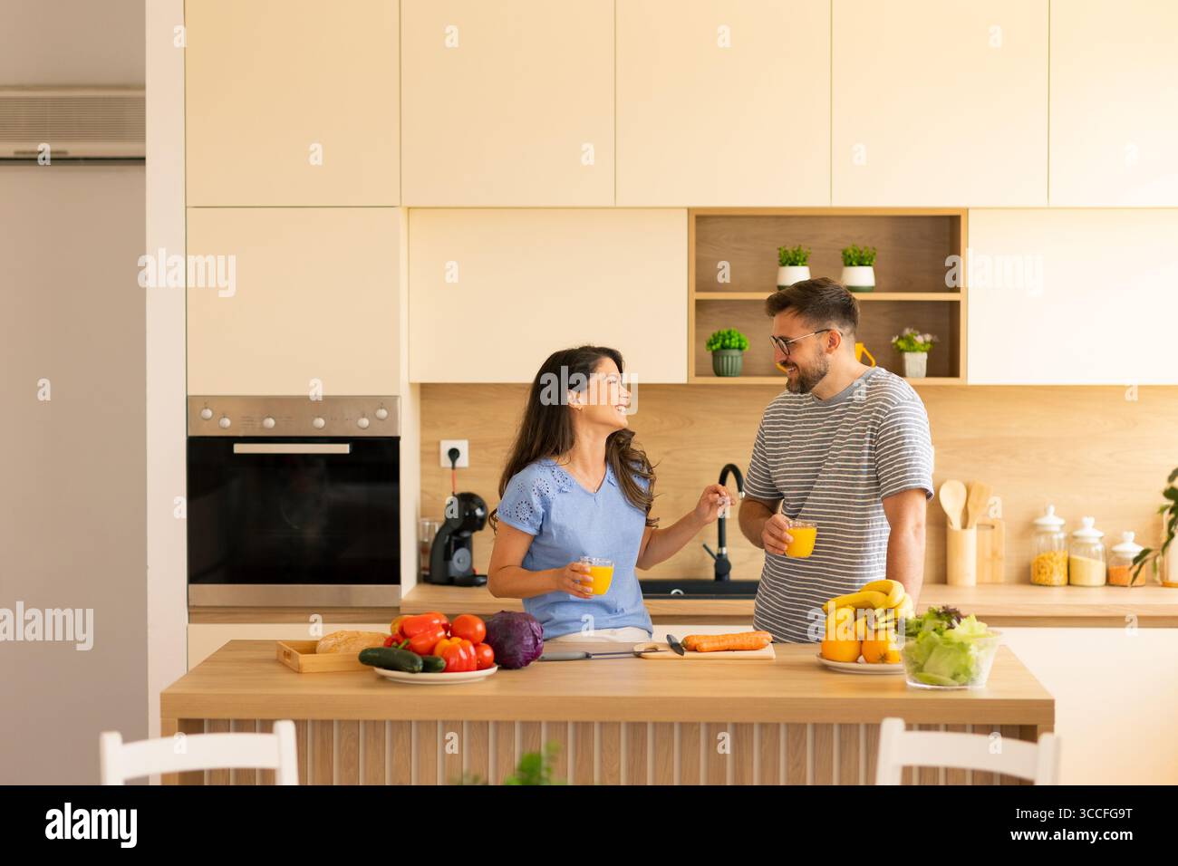 Due amici si godono un momento di allegria mentre preparano ingredienti freschi in una cucina elegante, assaporando il loro tempo insieme. Frutta e verdura brillanti Foto Stock