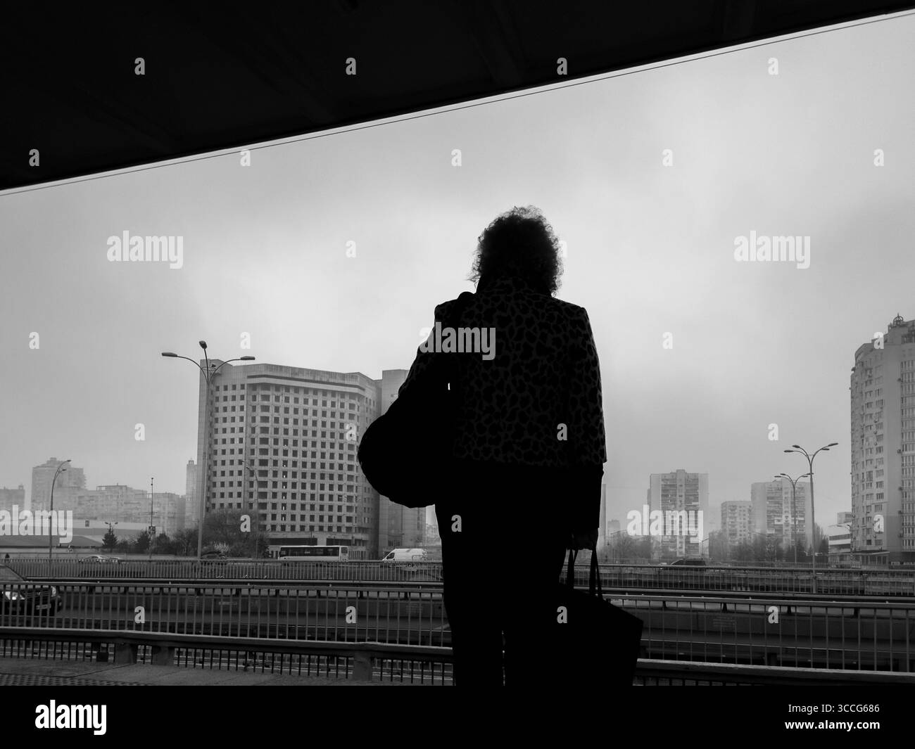 Kiev, Ucraina - 20 marzo 2025: Vista posteriore di una donna in piedi sulla metropolitana o sulla piattaforma ferroviaria che aspetta e guarda lo skyline della città con edifici e traffi Foto Stock