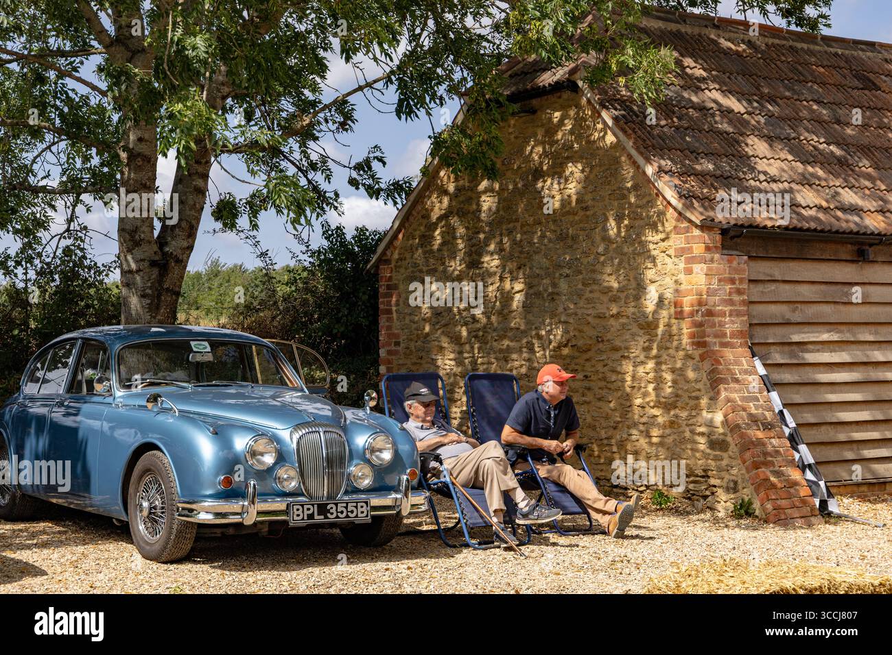 Due anziani si rilassano sulle sedie da campeggio accanto a una classica auto d'epoca blu parcheggiata da un rustico fienile di pietra al revival d'auto d'epoca dello stupido Post Inn Foto Stock