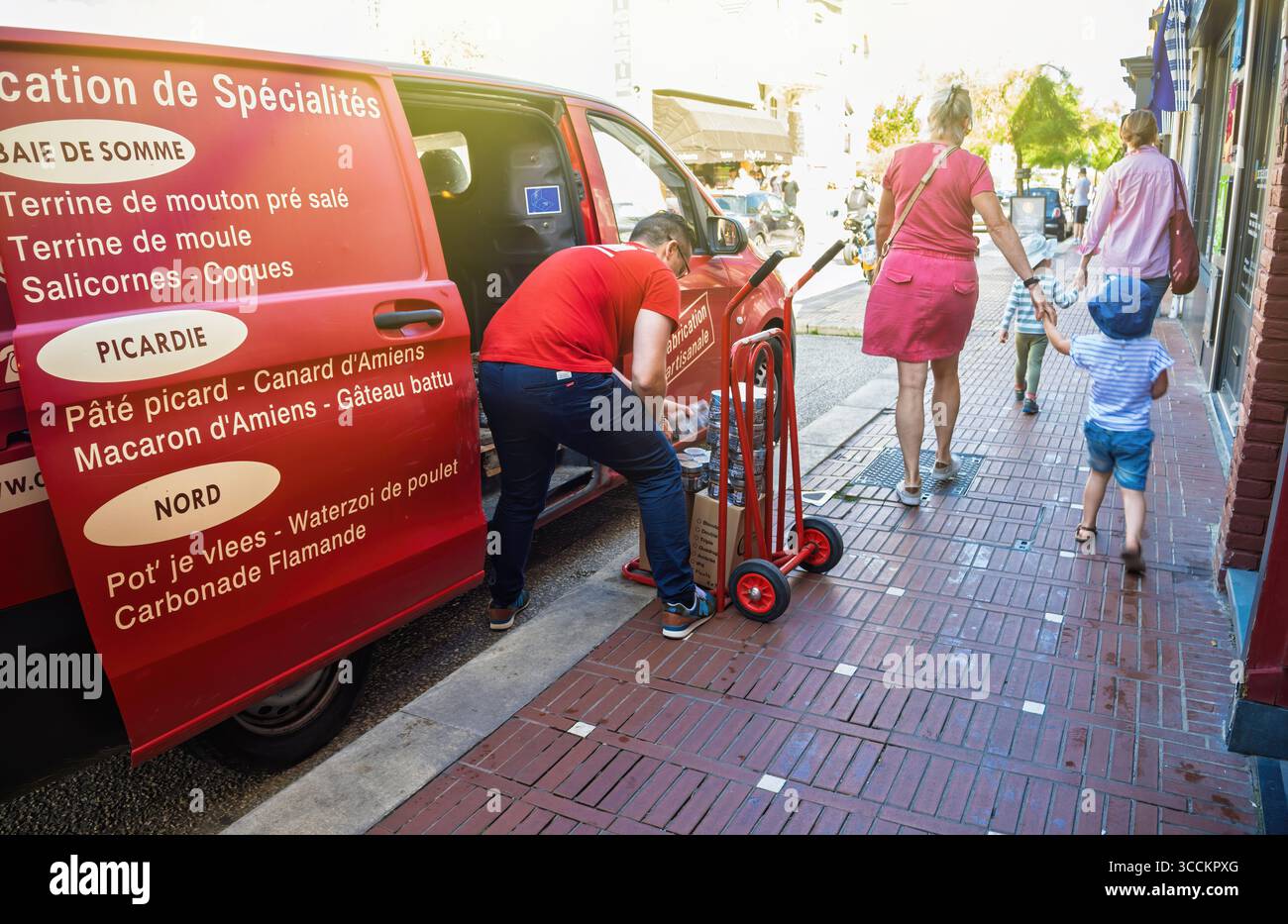 Le Touquet, Francia - 22 agosto 2023: Un furgone rosso che espone specialità locali è parcheggiato su un marciapiede di mattoni mentre un uomo scarica merci mentre le famiglie con Foto Stock