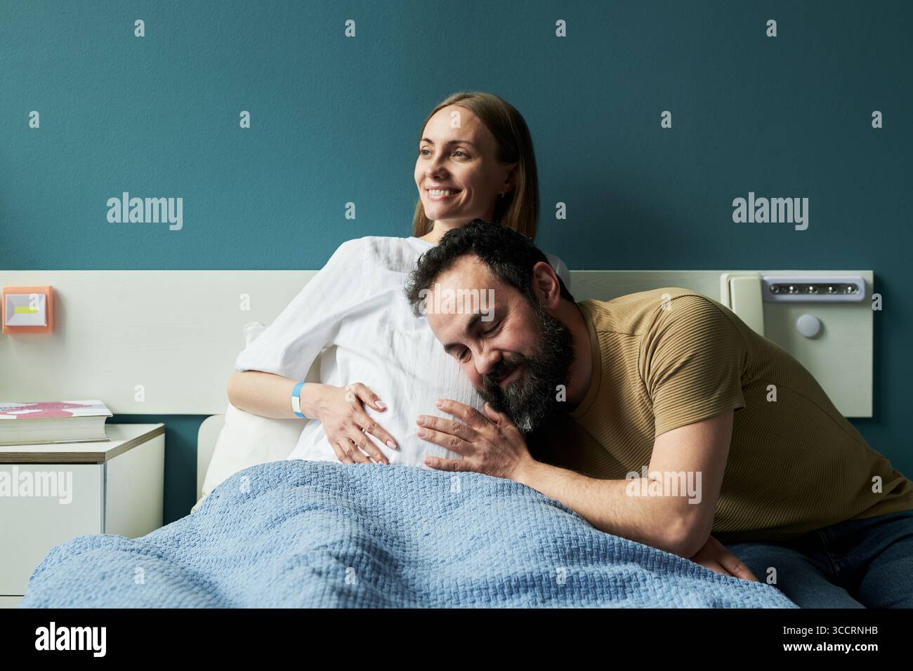 Caucasica incinta giovane donna adulta seduta nel letto d'ospedale sorridente mentre uomo appoggiato testa sulla sua pancia ascoltare e abbracciare lei in reparto maternità Foto Stock