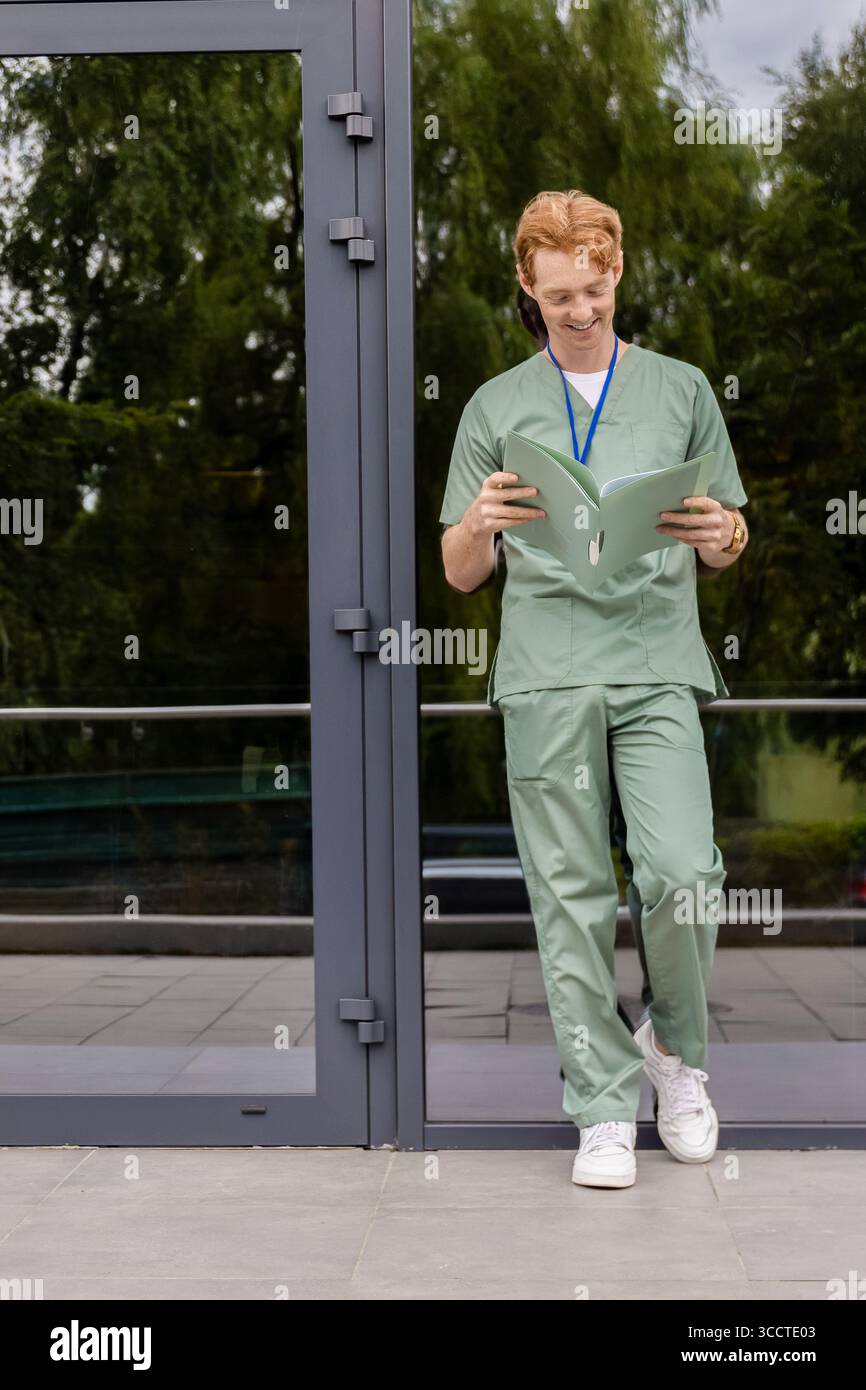 Medico caucasico in uniforme verde in piedi vicino alla facciata della clinica di vetro con badge e cartellino Foto Stock