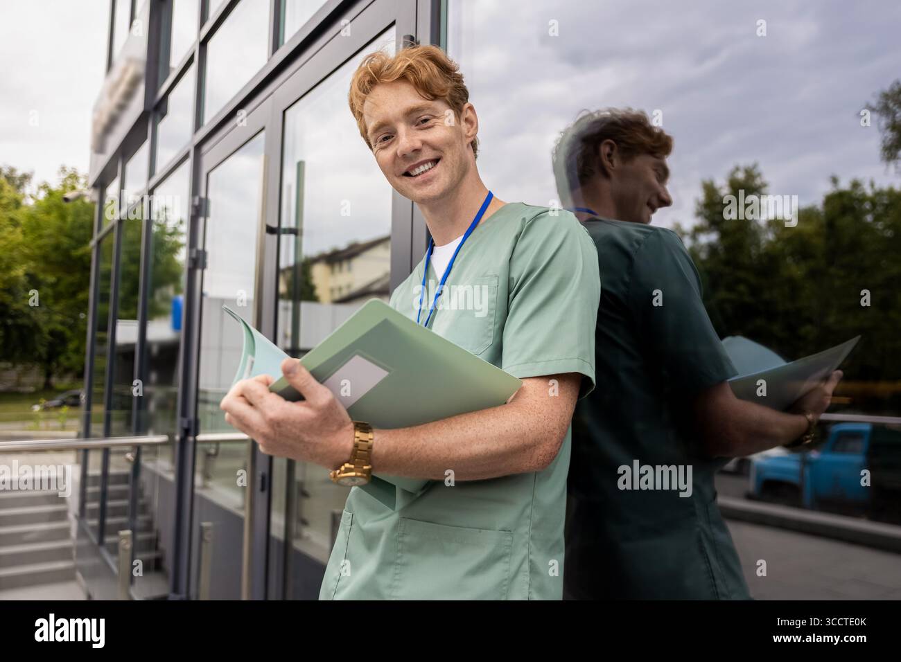 Zenzero medico in camice da laboratorio in piedi vicino alla clinica di vetro edificio tenendo cartella medica Foto Stock