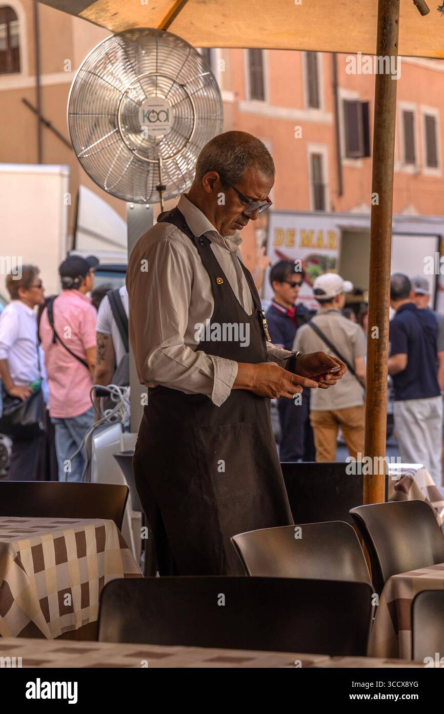 Un cameriere si trova di fronte al Pantheon, Roma, e pulisce i bicchieri da un ventilatore rinfrescante in un giorno rovente di luglio sotto i luminosi cieli estivi. Foto Stock