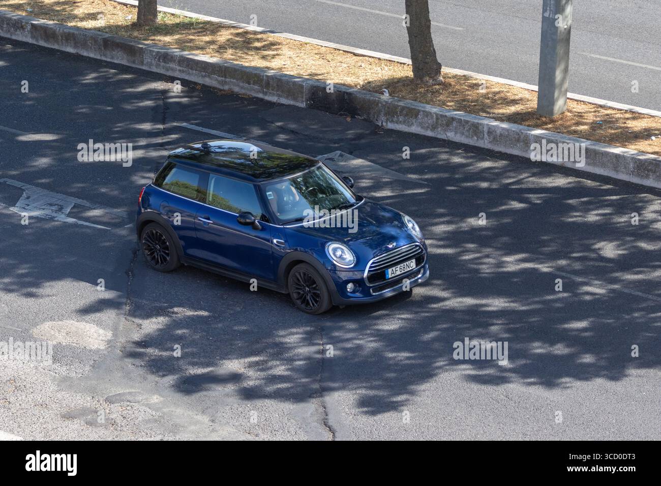 Moderna mini cooper blu che guida su una strada asfaltata vicino agli alberi, gettando ombre sul terreno Foto Stock