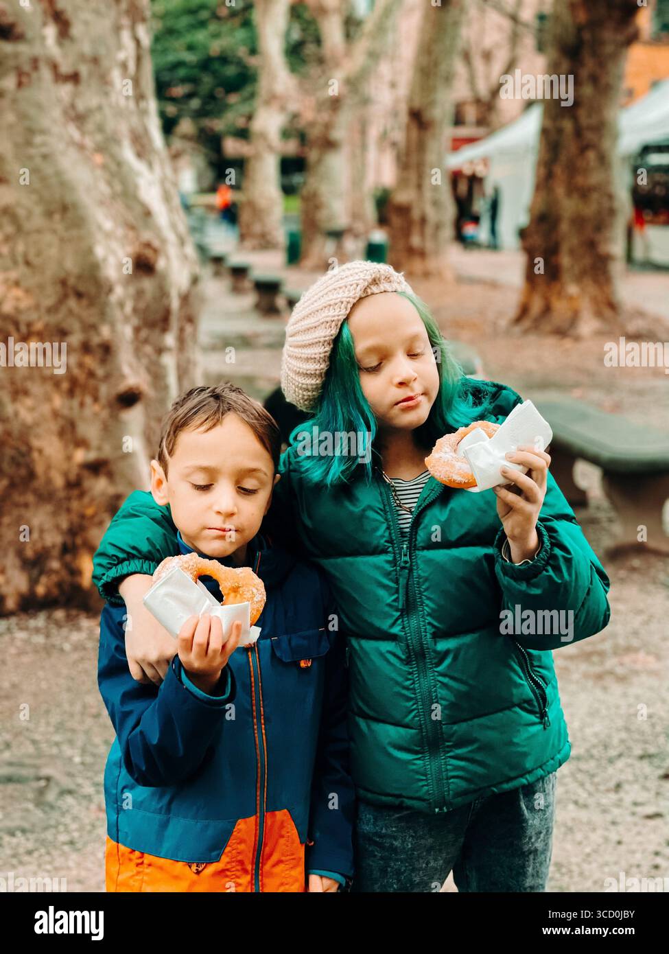 Due bambini sono felici di viziarsi con le ciambelle mentre si trovano in un parco panoramico. Indossati con calore per le giornate più fredde, condividono un momento di soddisfazione Foto Stock