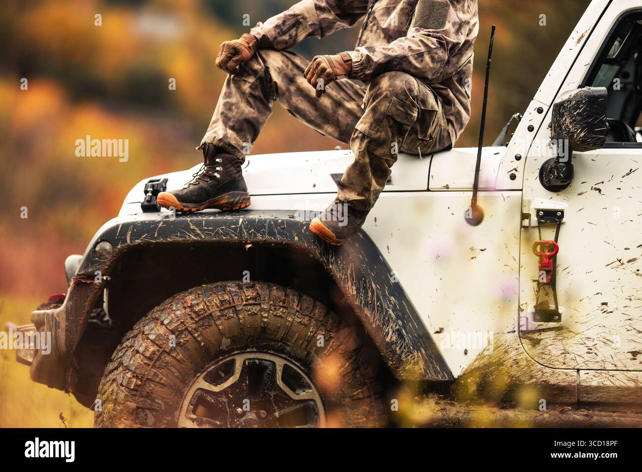 Un soldato riposa con una gamba sola su una jeep fangosa in un ambiente autunnale circondato da vegetazione colorata e natura, mostrando il brivido del fuoristrada explora Foto Stock