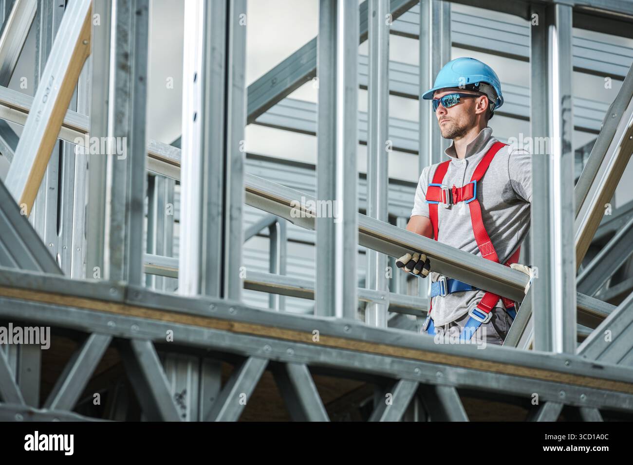 Un operaio edile in un elmetto solleva travi metalliche in un cantiere mentre indossa attrezzature di sicurezza. L'attività si svolge in un contesto moderno Foto Stock