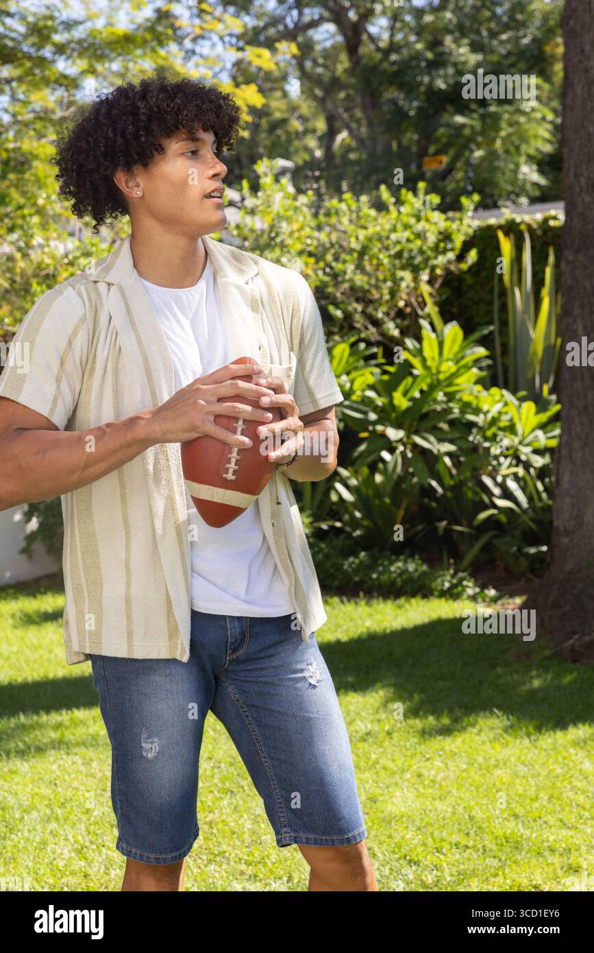 Uomo che indossa camicia a righe e pantaloncini corti su un prato verde nel cortile dell'hotel, che fa presa sul football americano Foto Stock