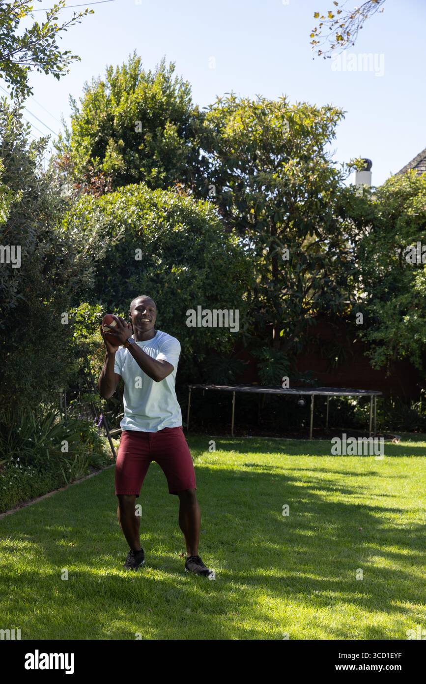Uomo afroamericano che cattura il football nel cortile vicino alla cornice del trampolino in pantaloncini rossi con camicia bianca Foto Stock