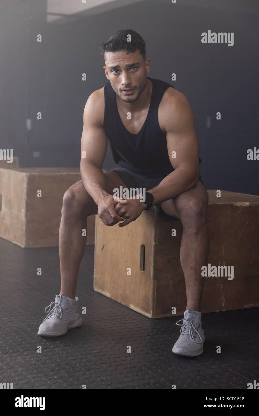 Uomo muscoloso seduto su una scatola plyometrica in legno su un tappetino in gomma in una palestra tenue con smartwatch Foto Stock