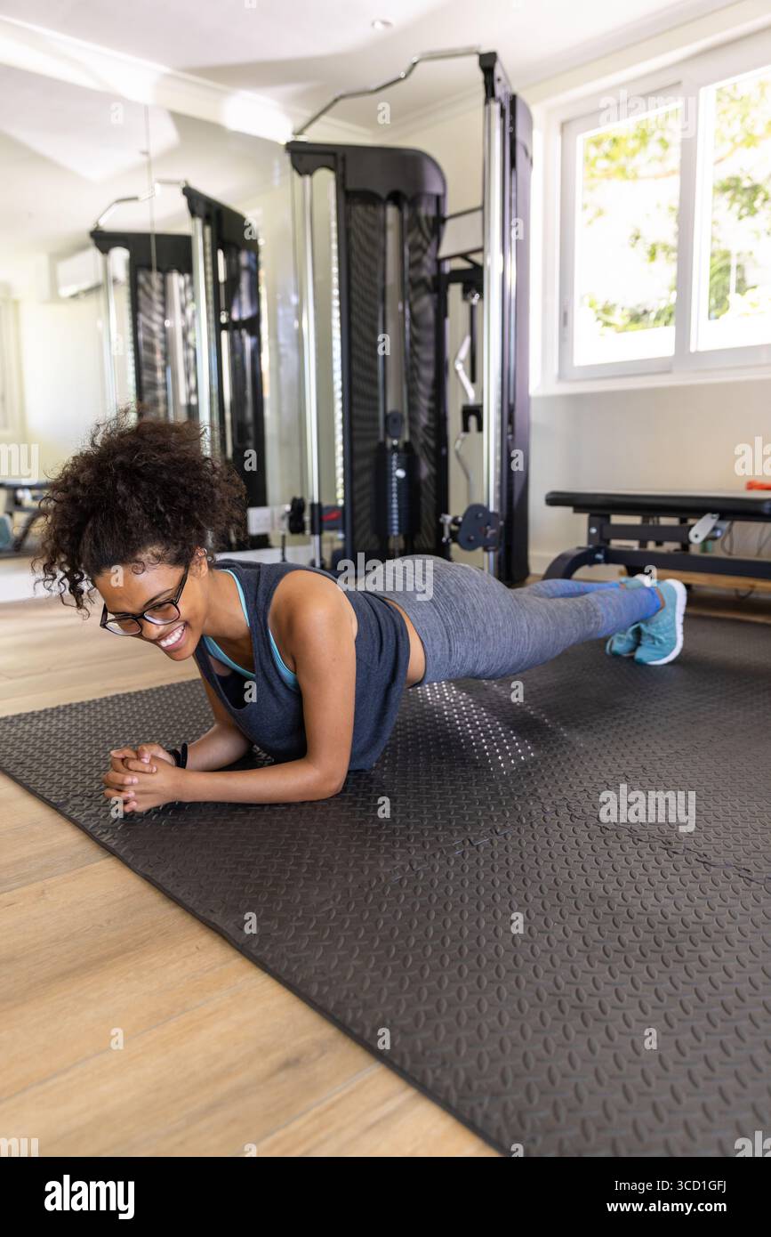 Donna afroamericana che indossa abbigliamento sportivo planking su un tappeto nero nella sala fitness con macchina per cavi Foto Stock