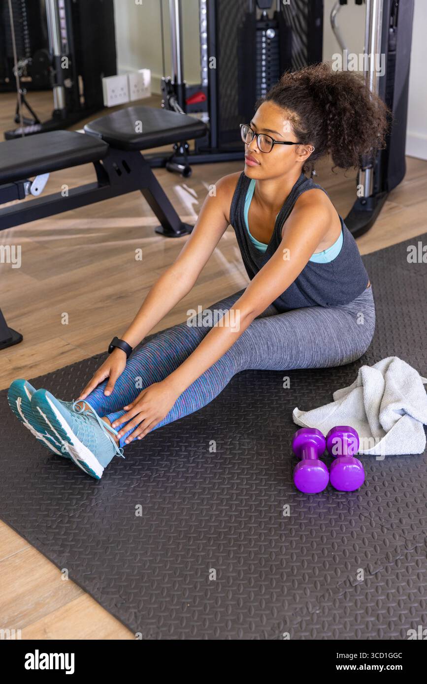 Donna afroamericana che si avvicina in avanti toccando le dita dei piedi sul tappetino per esercizi in palestra con manubri Foto Stock
