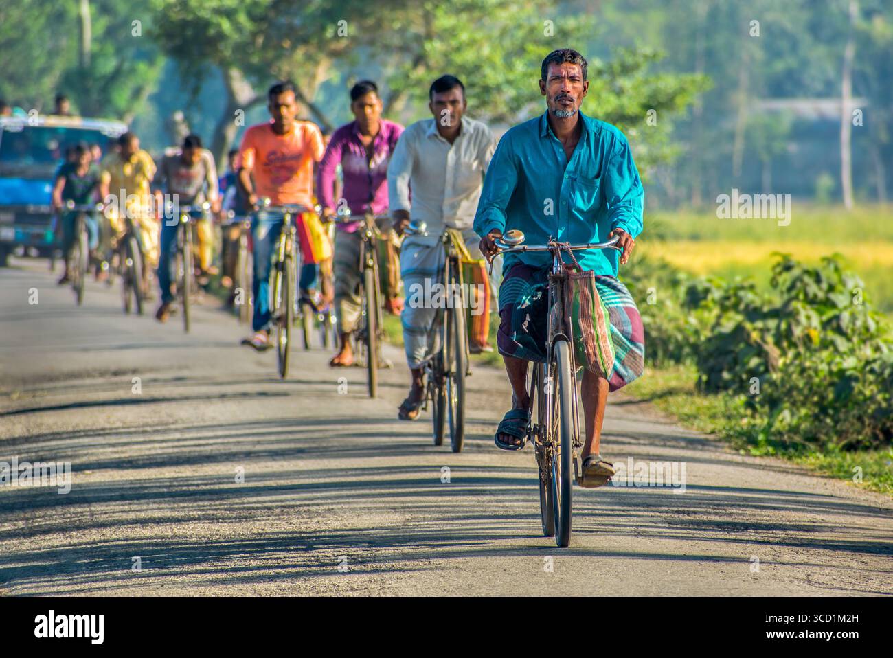 Bogura, Bangladesh - 7 novembre 2017: Veduta degli uomini in bicicletta lungo una strada soleggiata, con le loro camicie e i loro pranzi vivaci che si distinguono contro il verde fogliame. Foto Stock