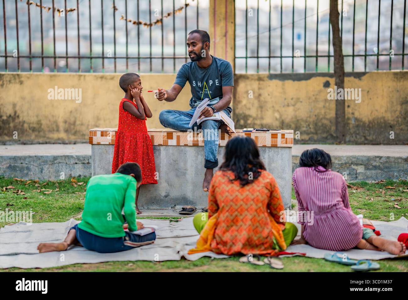 Bogura, Bangladesh - 5 aprile 2022: Vista di un uomo seduto su una panchina di cemento, che insegna ai bambini su un tappeto in una vivace scena all'aperto, incorniciata da una sfocatura Foto Stock