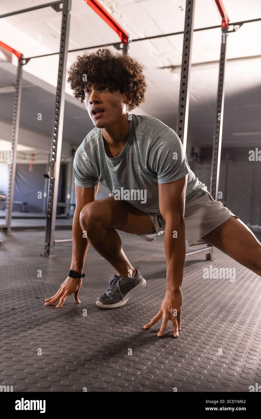 Uomo che esegue lo stretching laterale sul pavimento in gomma della palestra indossando il fitness tracker con uno squat rack in metallo Foto Stock