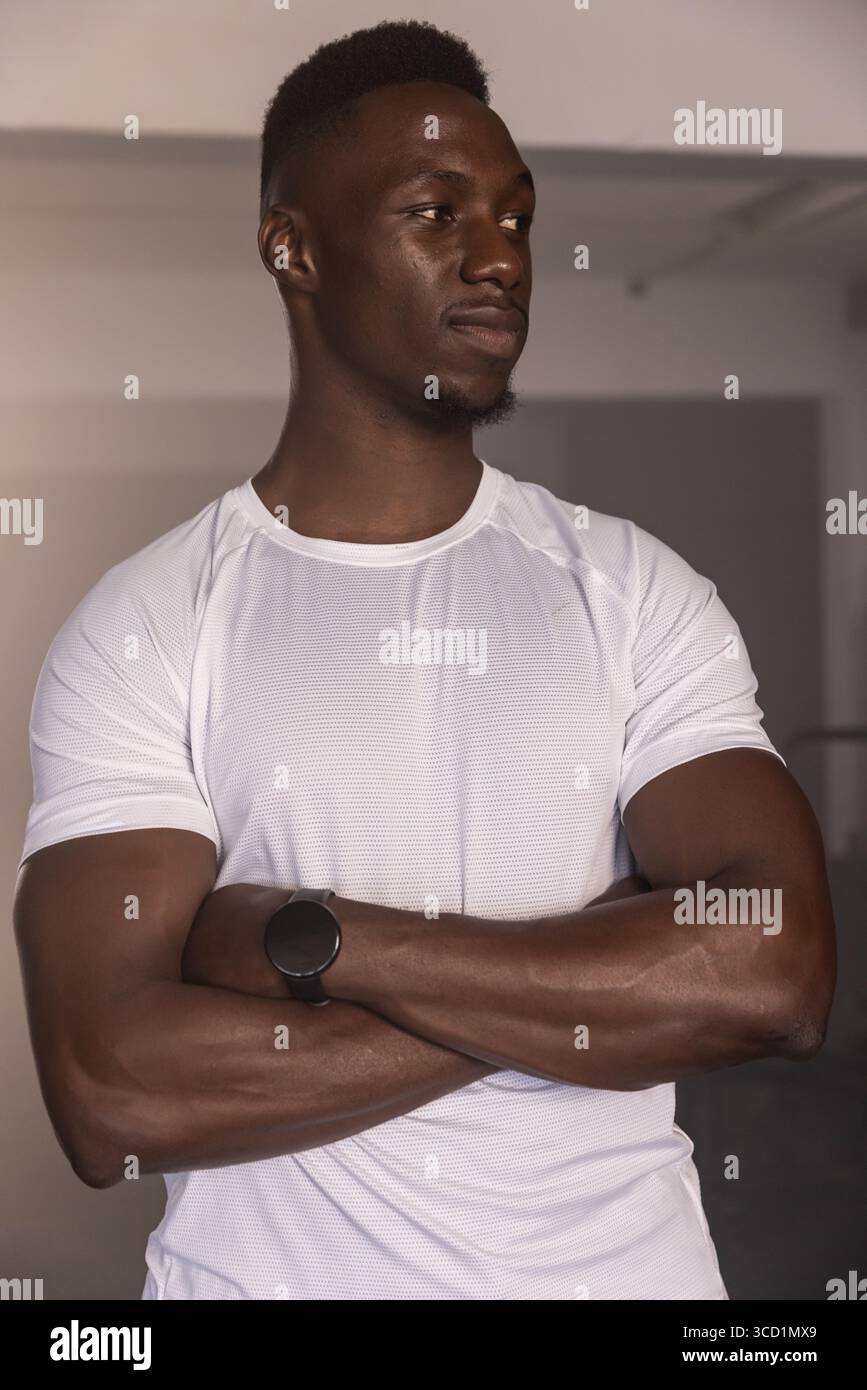 Uomo adulto afroamericano in piedi accanto alla parete grigia in palestra con t-shirt bianca e smartwatch nero Foto Stock