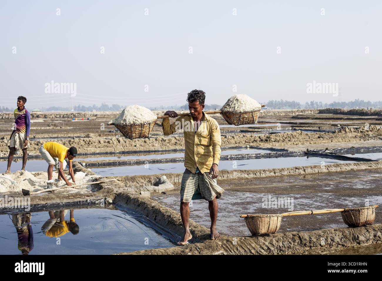 Banshakhali, Bangladesh - 06 febbraio 2023: Vista dei lavoratori che raccolgono sale nei campi scintillanti, le loro riflessioni rispecchiano il lavoro che si rompe sotto il vasto e pallido cielo. Foto Stock