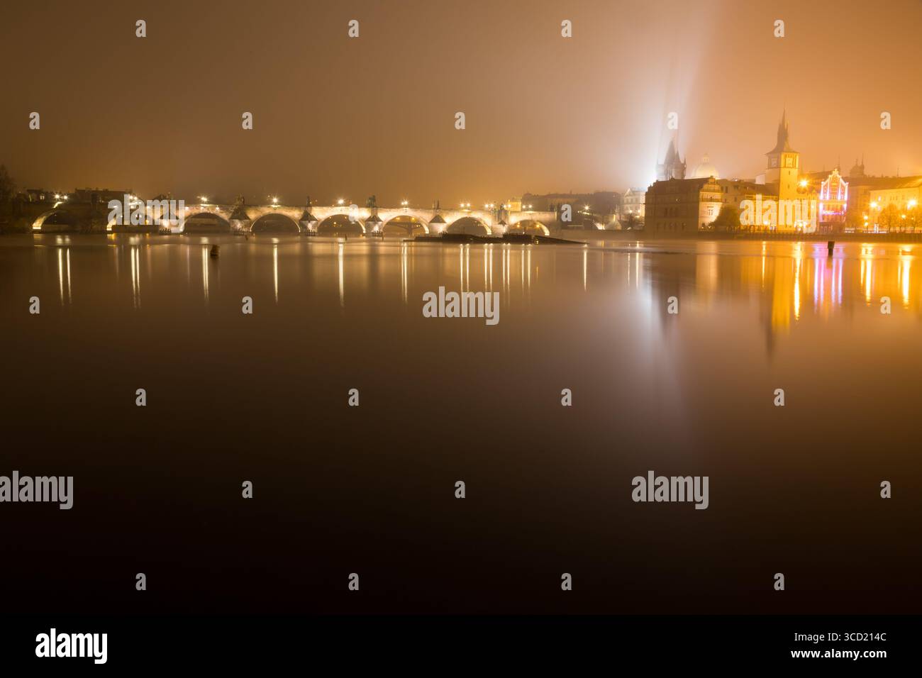 Il Ponte Carlo illuminato a Praga, che brilla di notte, si riflette nelle calme acque del fiume Moldava, le luci della città creano una scena dorata e nebbiosa Foto Stock