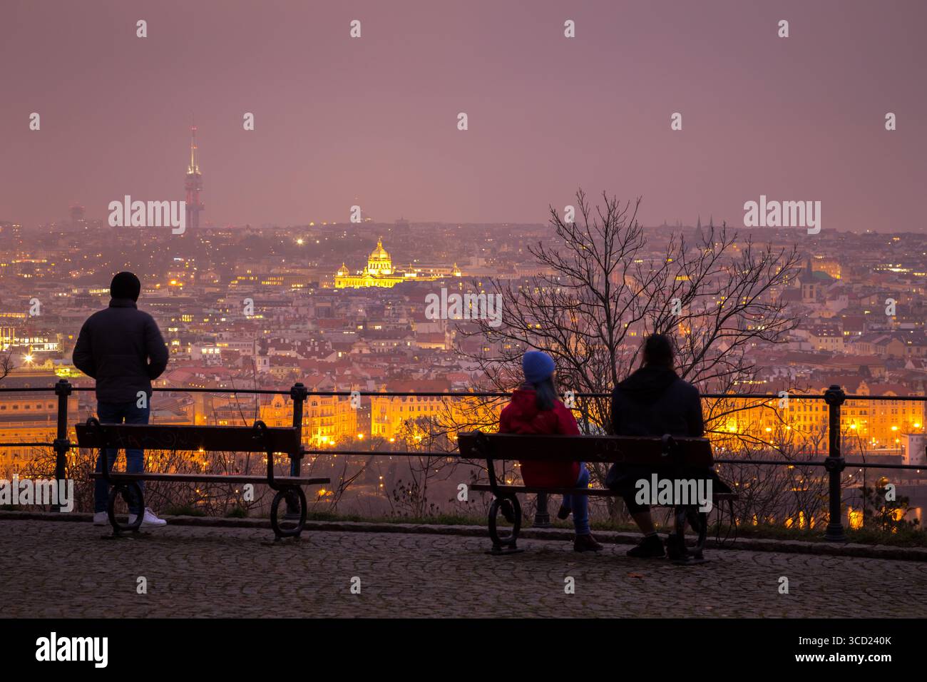 Persone che si affacciano sulla vista notturna di Praga illuminata in una serata nebbiosa sedute su una panchina Foto Stock