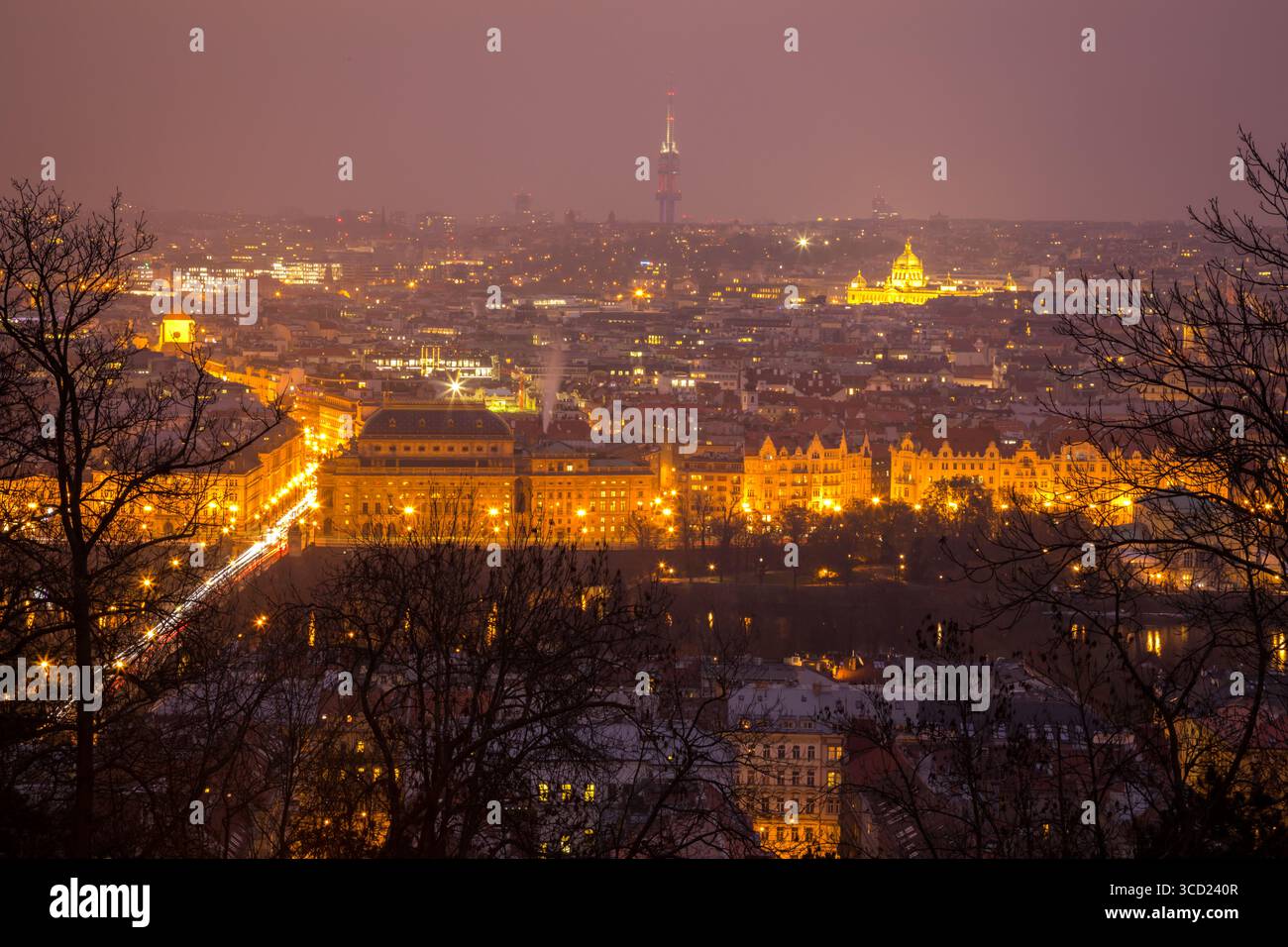 Vista notturna dei ponti e degli edifici illuminati di Praga che si illuminano contro un cielo notturno nebbioso, riflettendo le luci dorate della città nel fiume Foto Stock