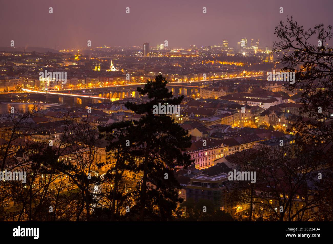 Vista notturna dei ponti e degli edifici illuminati di Praga che si illuminano contro un cielo notturno nebbioso, riflettendo le luci dorate della città nel fiume Foto Stock