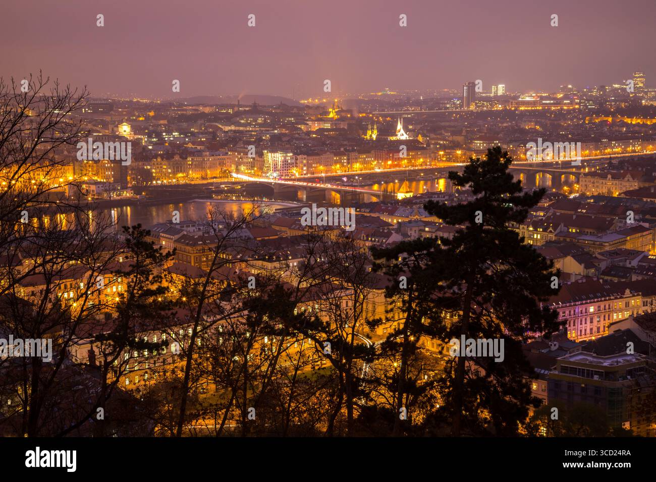 Vista notturna dei ponti e degli edifici illuminati di Praga che si illuminano contro un cielo notturno nebbioso, riflettendo le luci dorate della città nel fiume Foto Stock