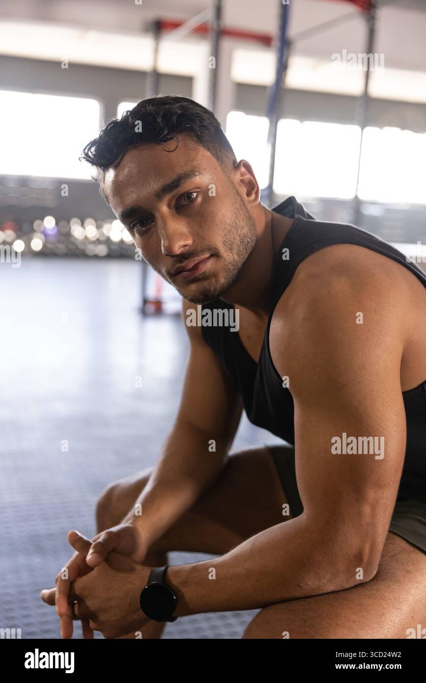 Uomo muscoloso seduto sul pavimento della palestra, che si piega in avanti e controlla lo smartwatch vicino al sistema di sollevamento Foto Stock